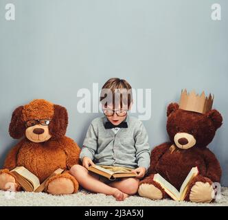 Getting some reading done. Studio shot of a smart little boy reading a book next to his teddy bears against a gray background. Stock Photo