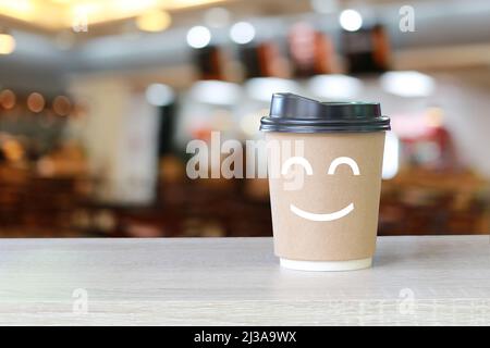 Paper cup of coffee on the table with a smile symbol on blur cafe background for design in your work. Stock Photo