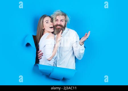 Couple through hole in blue paper, copy space for text or slogan. Discount and sale, people hole background. Surprised couple on blank paper hole Stock Photo