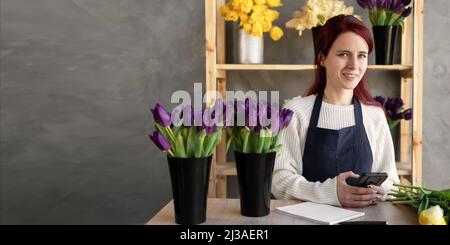 smiling florist in blue apron checks orders on mobile phone while sitting at a table with fresh flowers in glass vases in store. The concept of a Stock Photo