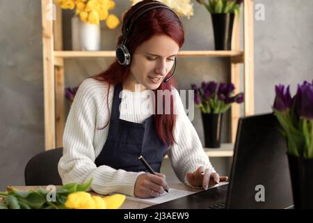 Young female florist in blue apron business owner using headset and computer while talking to customers online taking order online. Stock Photo