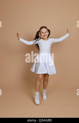 Young cute smiling girl with long dark hair in white dress, socks and gymnastics shoes dancing, spreading hands, raising leg, having fun on brown Stock Photo