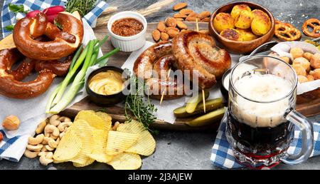 Octoberfest dinner table concept with grilled pork sausages, pretzel pastry, potatoes, salad, snacks, sauces, beer on dark background, top view. Stock Photo