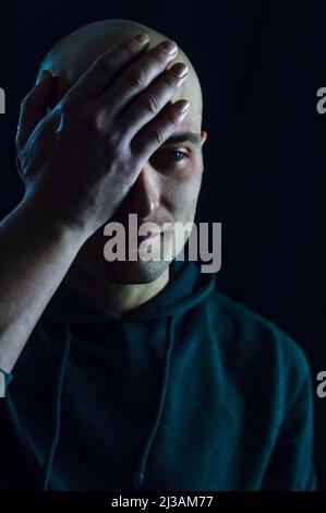 Portrait of a young bald man who partially covers his face with his hand on his forehead and looks sad Stock Photo