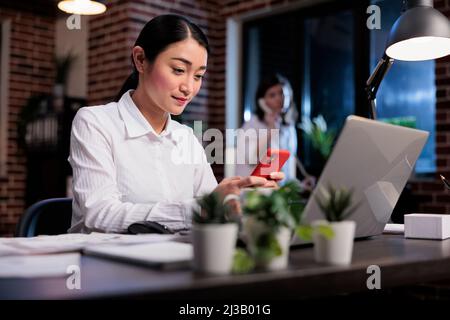 Businesswoman at work sending messages on social media platform using mobile phone. Happy smiling asian office worker sitting in office workspace while texting somebody on modern smartphone device. Stock Photo