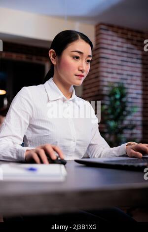 Portrait shot of confident businesswoman at night sitting in office workspace while working on laptop. Management department leader analyzing financial charts and graphs while checking documentation. Stock Photo