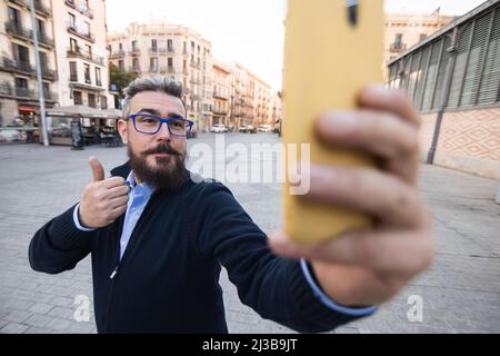 Man showing thumb up while taking a selfie with a mobile phone outdoors on the street. Stock Photo