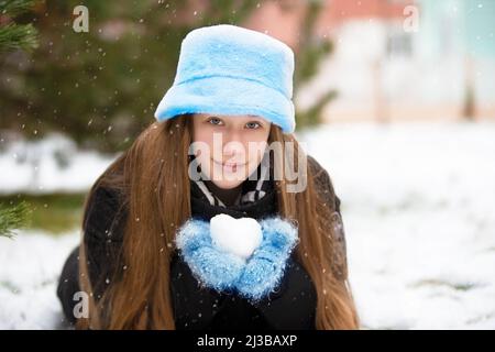 A beautiful girl in a fur hat and mittens holds a snowball in her hands. Stock Photo