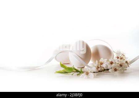 Three white eggs for Easter, a ribbon and with a branch of wild cherry flowers on a light background with copy space, holiday greeting card, selected Stock Photo