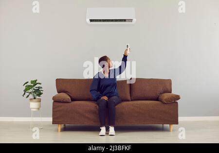 Young african woman switching on air conditioner with remote control at home. AC system concept. Stock Photo