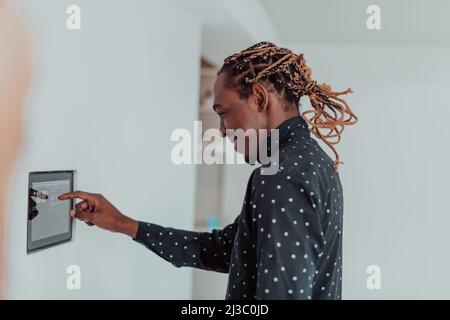 Smiling African American man using modern smart home system, controller on wall, positive young man switching temperature on thermostat or activating Stock Photo