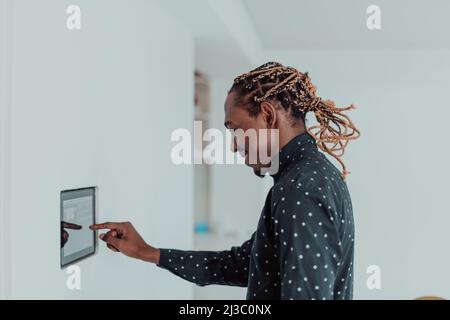 Smiling African American man using modern smart home system, controller on wall, positive young man switching temperature on thermostat or activating Stock Photo