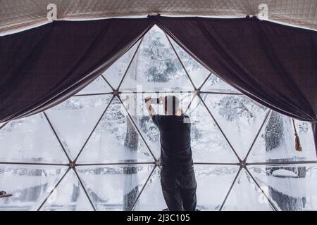 A man takes pictures on his smartphone of a view of a snow-covered forest from a glamping dome tent. Stock Photo