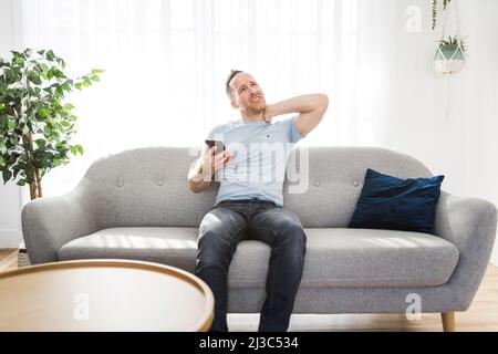 man sitting on couch touching massaging neck he suffering from neck pain ache after using phone Stock Photo