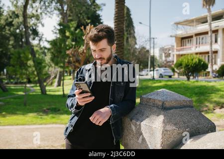 Young man wearing blue jean jacket leaning to a decorative stone in a park. Looking to phone scrolling, messaging or watching video. Stock Photo