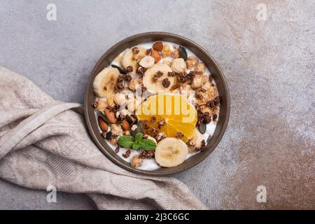 Top view of granola with yogurt, fresh banana, orange, almonds and hazelnuts, pumpkin seeds for healthy breakfast on grey concrete background Stock Photo