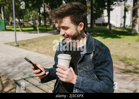 Smiling, young man holding coffee cup smiling at his phone while sitting on bench, a pleasant moment and enjoy Stock Photo