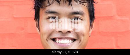 Horizontal banner or header with portrait of handsome young asian man standing outdoors against a red wall background in city and looking at camera - Stock Photo