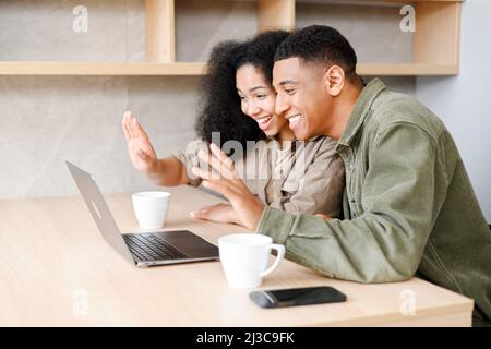 Multiracial couple using laptop for video call while sitting at the table in cozy kitchen. Cheerful spouses waving into webcam, involved virtual meeting with family. Online communication concept Stock Photo