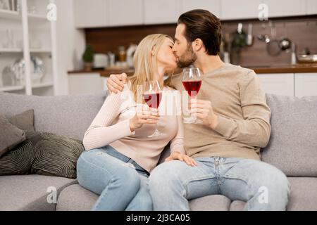 Happy marriage anniversary. Cheerful spouses celebrating anniversary at home, sitting on the sofa and holding glasses with wine, enjoying time together, kissing Stock Photo
