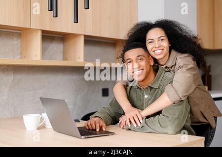 Biracial woman hugging man from behind. Mixed ethnicity couple relaxing in new apartment at home while browsing internet on laptop. Show love everyday in the simplest of ways concept Stock Photo