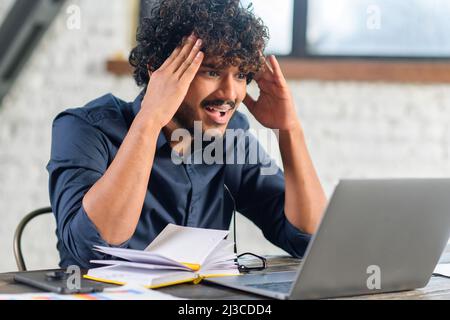 Tired Indian male office employee screaming while looking at the laptop screen, feeling boring and angry. Bored eastern man in smart casual shirt sitting at the desk Stock Photo