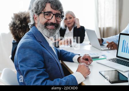 Senior business man smiling at camera while working with colleagues inside bank office - Focus on face Stock Photo