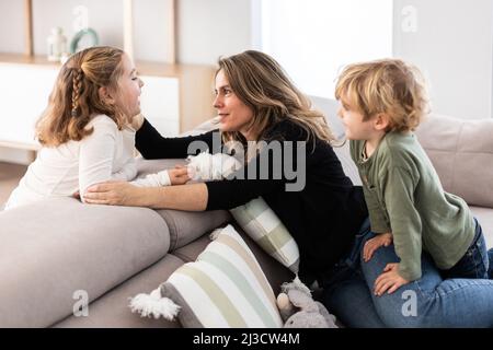 Caring mother and daughter looking at each other while sitting on comfortable couch with cute son in light living room Stock Photo