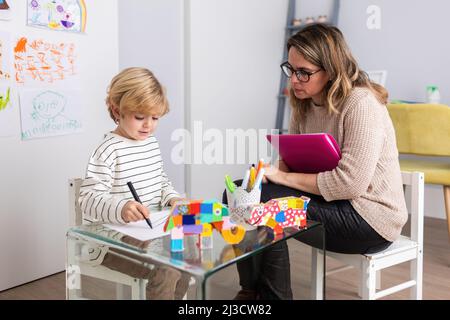 Focused female psychologist with folders sitting near boy drawing on paper at table with toys during appointment in light room Stock Photo