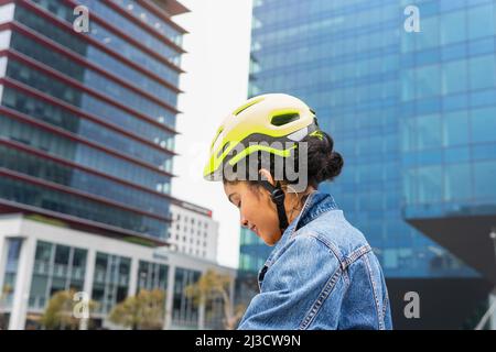 Side view of s smiling young Colombian female in stylish denim outfit wearing protective helmet before riding bike on street of modern city Stock Photo