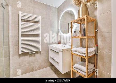 Modern bathroom with towels on wooden shelves and ceramic sink under illuminated mirror Stock Photo