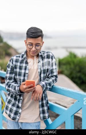 Happy ethnic young male in casual clothes and glasses smiling and browsing social media on cellphone while leaning on blue railing of bridge on summer Stock Photo