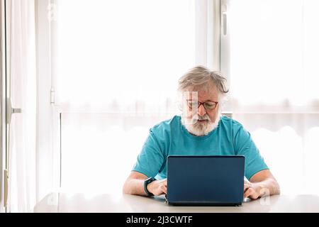 Focused senior male with eyeglass browsing on laptop sitting on a table at home Stock Photo