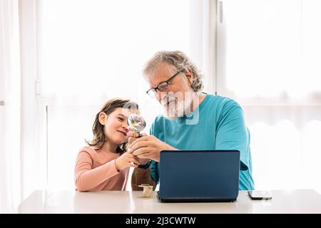 Positive grandfather with cute granddaughter looking at white chamomile through magnifying glass while sitting at table with laptop in room Stock Photo