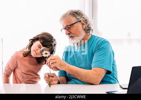 Content bearded grandfather with positive girl looking at white chamomile through magnifying glass while studying botany in light room at home Stock Photo