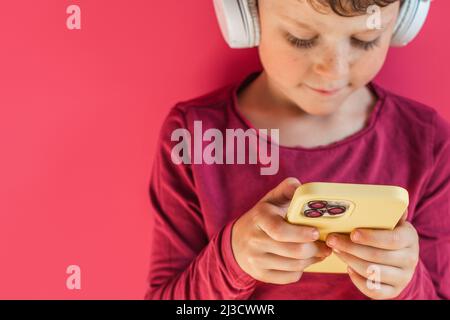 Serious boy listening to music in headphones and browsing modern cellphone in studio against pink background and looking at screen Stock Photo