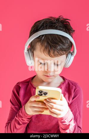 Serious boy listening to music in headphones and browsing modern cellphone in studio against pink background and looking at screen Stock Photo