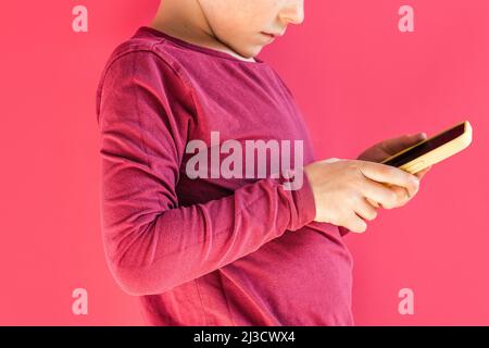 Side view of crop unrecognizable digitally addicted boy browsing cellphone in bright studio against pink background Stock Photo