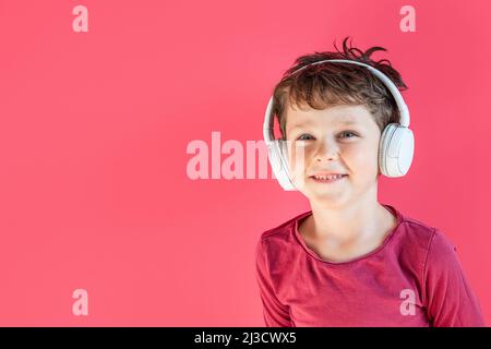 Happy kid with wireless headphones while listening to music in leisure time in studio against pink background and looking at camera Stock Photo