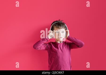 Optimistic kid keeping hands on wireless headphones while listening to music in leisure time in studio against pink background and looking at camera Stock Photo