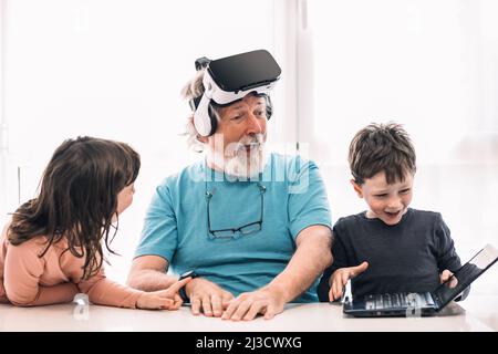 Bearded grandfather in modern VR headset playing videogame on laptop while sitting at table near cute children in light room Stock Photo