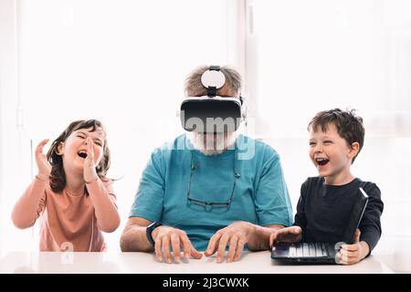 Bearded grandfather in modern VR headset playing videogame on laptop while sitting at table near cute children in light room Stock Photo