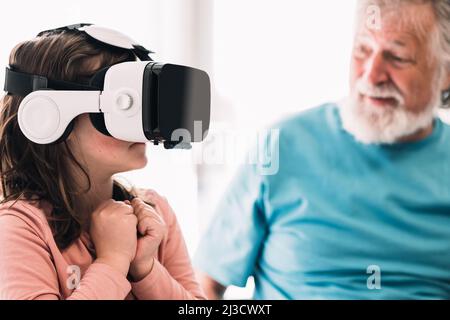 Cute girl in modern VR goggles exploring cyberspace while sitting near crop content bearded grandfather in light room at home Stock Photo