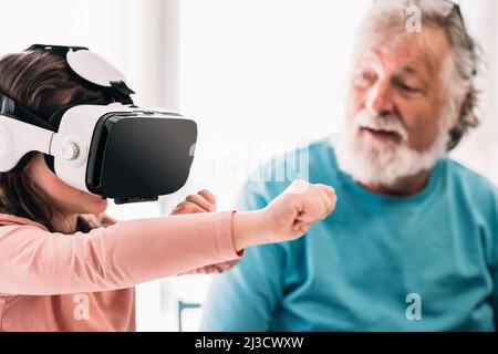 Cute girl in modern VR goggles exploring cyberspace while sitting near crop content bearded grandfather in light room at home Stock Photo