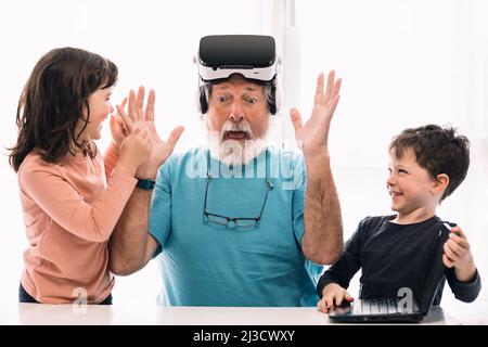 Amazed bearded grandfather in VR headset sitting near positive children while exploring cyberspace at table with laptop in light room Stock Photo
