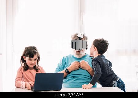 Scared bearded grandfather in VR headset sitting near positive children while exploring cyberspace at table with laptop in light room Stock Photo
