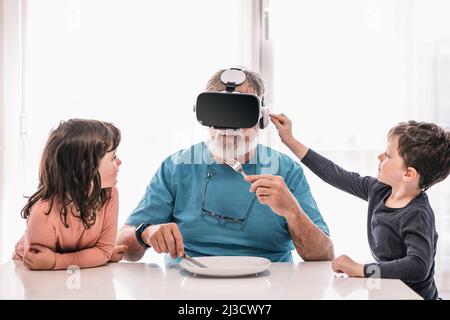 Senior male in modern VR goggles sitting at table with tableware while eating and experiencing virtual reality with his grandchildren Stock Photo