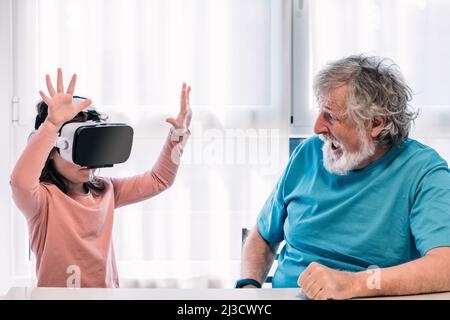 Cute girl in modern VR goggles exploring cyberspace with raised arms while sitting near bearded grandfather in light room at home Stock Photo