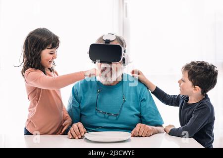 Positive children pretending to feed bearded grandfather in VR goggles exploring virtual reality at table in light room at home Stock Photo