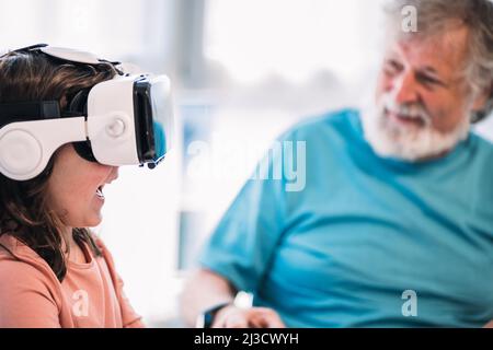 Cute girl in modern VR goggles exploring cyberspace while sitting near crop content bearded grandfather in light room at home Stock Photo